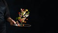 The chef preparing tasty dish with meat and mix of vegetables in a griddle, on a black background. Backstage of preparing