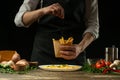 Chef salting in the envelope french fries ready, on the background with vegetables. Cooking tasty but harmful food. Background for Royalty Free Stock Photo