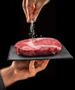 Chef is salting or seasoning raw ribeye steak laying on graphite serving board. Black background