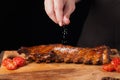 The chef sprinkles salt in ready to eat pork ribs, lying on an old wooden table. A man prepares a snack to beer on a black backgro