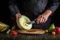 A chef in uniform peels a ripe melon with a knife before slicing it for table service in a restaurant. Fruit diet concept on a Royalty Free Stock Photo