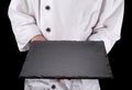 Chef in white uniform is holding an empty black slate serving plate. Black background Royalty Free Stock Photo