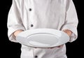 Chef in white uniform is holding an empty white round ceramic serving plate. Black background Royalty Free Stock Photo