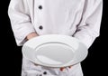 Chef in white uniform is holding an empty white round ceramic serving plate. Black background Royalty Free Stock Photo