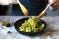A chef with a wooden spatula and spoon mixes broccoli in a pan.