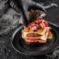 Chefs hand in black glove cooking the black burger with beaf steak, lettuce, tomatoes, slice of cheese, ham, pastrami and sauce on Royalty Free Stock Photo