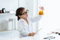 In chemistry classroom with many laboratory tools on shelves and table. A little Asian girl with glasses looking yellow chemical