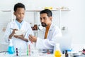 In chemistry classroom with many laboratory tools on table. A young African boy and male teacher in white lab coat doing