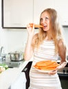 Cherful ordinary pregnant woman eating carrots at home interior