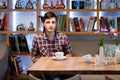 Handsome young man in checkered shirt sitting at wooden table and looking at camera in front of    shelves with books Royalty Free Stock Photo