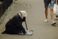 An elderly woman in a scarf beg for money, kneeling on a city street. A guy in shorts walks past