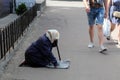 An elderly woman in a scarf beg for money, kneeling on a city street. A guy in shorts walks past