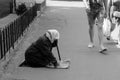 An elderly woman in a scarf beg for money, kneeling on a city street. A guy in shorts walks past