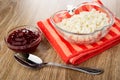 Cherry jam in bowl, bowl with grained cottage cheese on red napkin, spoon on wooden table