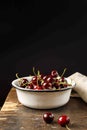 Cherry in a white metal bowl on a dark wooden table, vertical on a black background with space. Royalty Free Stock Photo