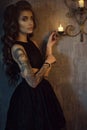 Chic young tattooed woman with luxurious long wavy silky hair standing in the dark shabby room at the burning candles