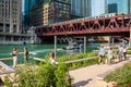 Chicago River scene of police tugboat on patrol, tourists and commuters walking & taking selfies, and el train crossing elevated