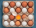 Chicken eggs in the carton box. Chicken egg is half broken among other eggs, eggshell and chicken feather near. Top view Royalty Free Stock Photo