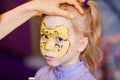 Child animator, artist`s hand draws face painting to little girl. Child with funny face painting. Painter makes yellow leopard at