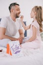 Child applying lipstick on fathers lips near gift box and fathers day greeting card on bedding