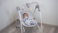 The child in the automatic swing cradle lies and eats from the bottle. The boy lies in a cradle at home.