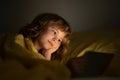 Child in bed under a blanket plays on a tablet in a game in the dark. The childs face is illuminated by a tablet. Child Royalty Free Stock Photo