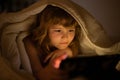 Child in bed under a blanket plays on a tablet in a game in the dark. The childs face is illuminated by a tablet. Child Royalty Free Stock Photo