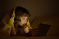 Child in bed under a blanket plays on a tablet in a game in the dark. The childs face is illuminated by a tablet. Child