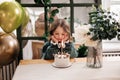 Child birthday party. Adorable little girl sits by the table with birthday cake decorated flowers and burn candle, makes