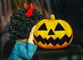 A child in a black wig with red horns hugs a pumpkin at a Halloween party. Adorable little girl hugs a big pumpkin on a pumpkin pa Royalty Free Stock Photo