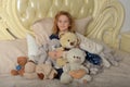 Child blonde in a blue dress sitting on the bed with many toy bears Royalty Free Stock Photo