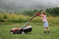 A child in boots in the form of a game mows grass with a lawnmower in the yard against the background of mountains Royalty Free Stock Photo