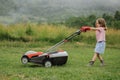 A child in boots in the form of a game mows grass with a lawnmower in the yard against the background of mountains Royalty Free Stock Photo