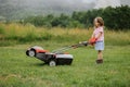 A child in boots in the form of a game mows grass with a lawnmower in the yard against the background of mountains Royalty Free Stock Photo