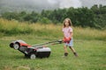 A child in boots in the form of a game mows grass with a lawnmower in the yard against the background of mountains Royalty Free Stock Photo