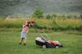 A child in boots in the form of a game mows grass with a lawnmower in the yard against the background of mountains Royalty Free Stock Photo