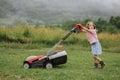 A child in boots in the form of a game mows grass with a lawnmower in the yard against the background of mountains Royalty Free Stock Photo