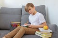 Child boy hardly working on his homework, studying online at home with books and laptop, sitting on grey sofa in modern interior, Royalty Free Stock Photo