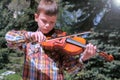 Portrait of child boy is playing the violin standing in park on pine background.