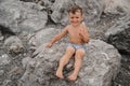 The child boy is sitting, smiling and sunbathing on large boulders, on the beach near the sea. Royalty Free Stock Photo