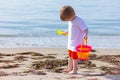 Child boy with toy bucket and shovel in hands standing on sea beach and looking at algae underfoot Royalty Free Stock Photo