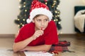 Child boy wearing Santa hat writing Christmas letter to Santa Claus at home near the Christmas tree Royalty Free Stock Photo