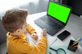 A child, a boy in a yellow sweater, is sitting at a table at home looking in a beech laptop with a green screen. The concept of