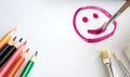 A child with a brush and raspberry paint draws a happy smile or smiling face on a sheet of white paper, on the sheet are colored