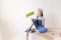 A child Builder sits on a construction ladder in an apartment with white walls and a roller in his hands and shows a thumbs up, a Royalty Free Stock Photo