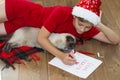 Child with a cat writing Christmas letter to Santa Claus. Boy wearing a Santa hat at home near the Christmas tree Royalty Free Stock Photo