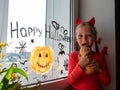 Child chewing pumpkin painting on window preparing to celebrate Halloween. Little girl draws decorates room