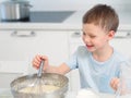 Child cooks food. Caucasian boy sits at table in kitchen and kneads dough in cup with hand whisk. Cheerful and positive Royalty Free Stock Photo