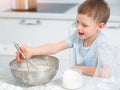 Child cooks food. Caucasian boy sits at table in kitchen and kneads dough in cup with hand whisk. Cheerful and positive Royalty Free Stock Photo