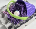A polkadot purple Easter basket with lime green handle rests on an open empty egg carton. See one egg and paper grass.